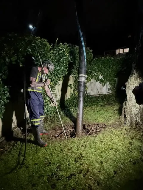A technician clearing a line after dark, hose fed down through a cleanout in the yard.